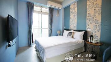 A hotel room with a large bed, a flat-screen TV, a bedside table with a lamp, and a window with curtains.