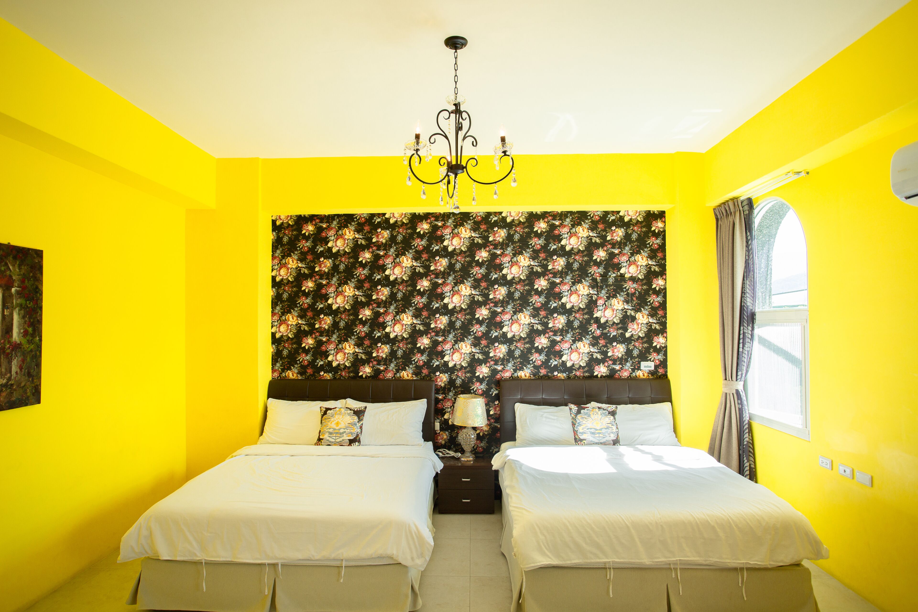 Una habitación con dos camas, una araña, papel tapiz floral y un ventanal con cortinas.