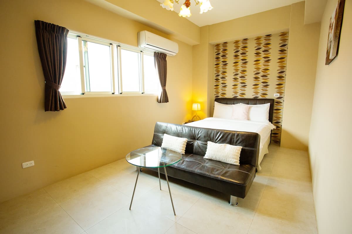 A hotel room with a bed, a sofa, a glass table, a wall-mounted air conditioner, a patterned wallpaper, and a wall socket.