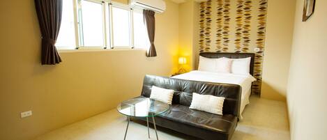 A hotel room with a bed, a sofa, a glass table, a wall-mounted air conditioner, a patterned wallpaper, and a wall socket.