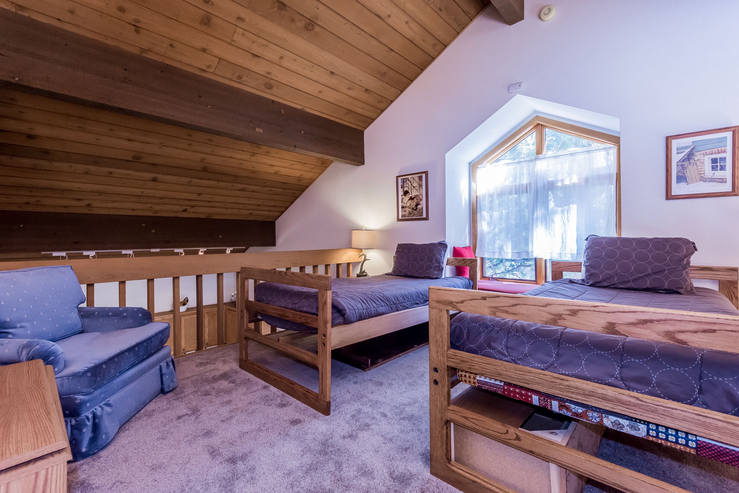 A cozy loft bedroom with a sloped ceiling, a wooden bed frame, a blue sofa, and a window with a view of trees.