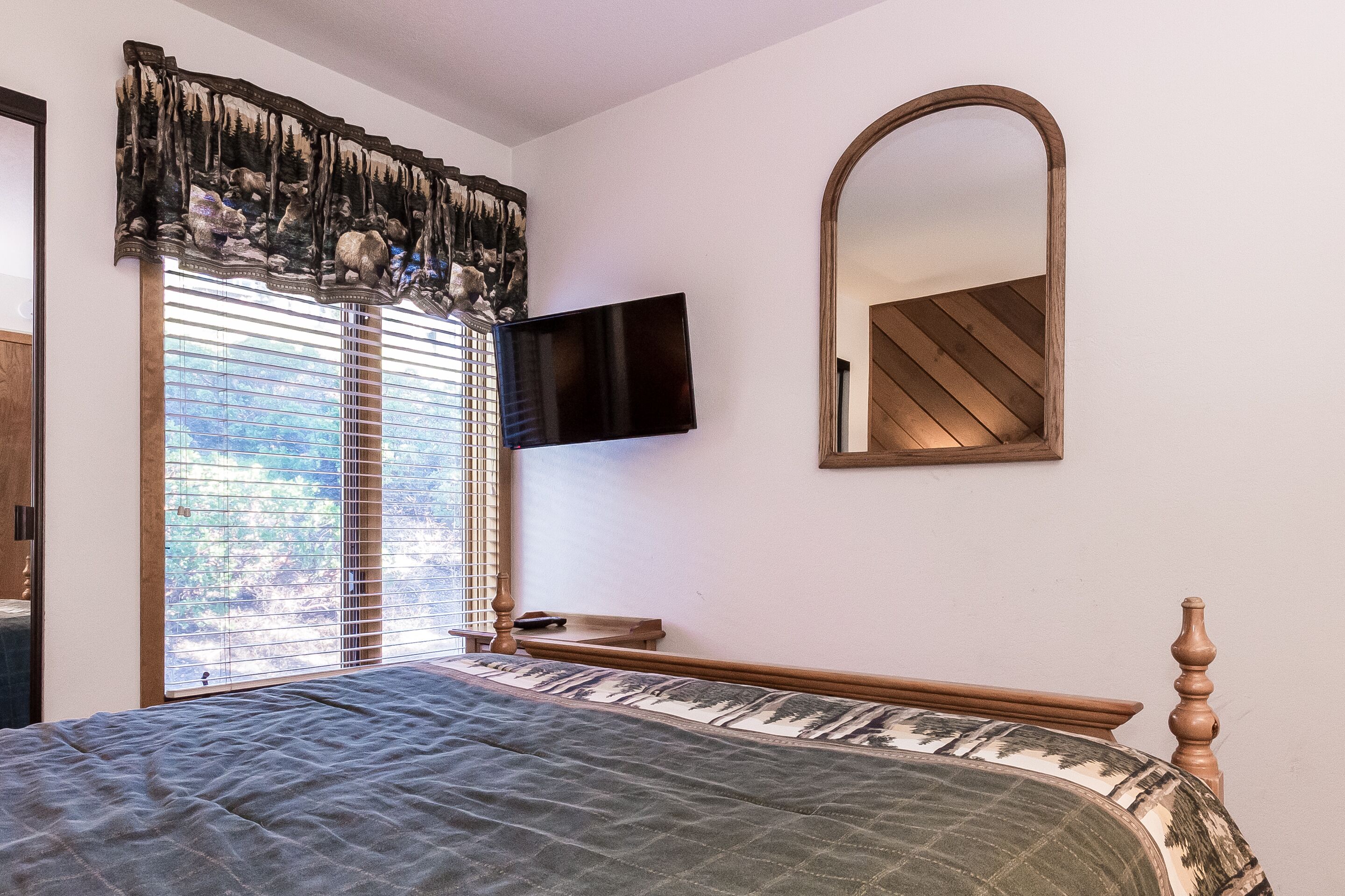 Un dormitorio con cama, televisor montado en la pared, un espejo grande y una ventana con persianas.