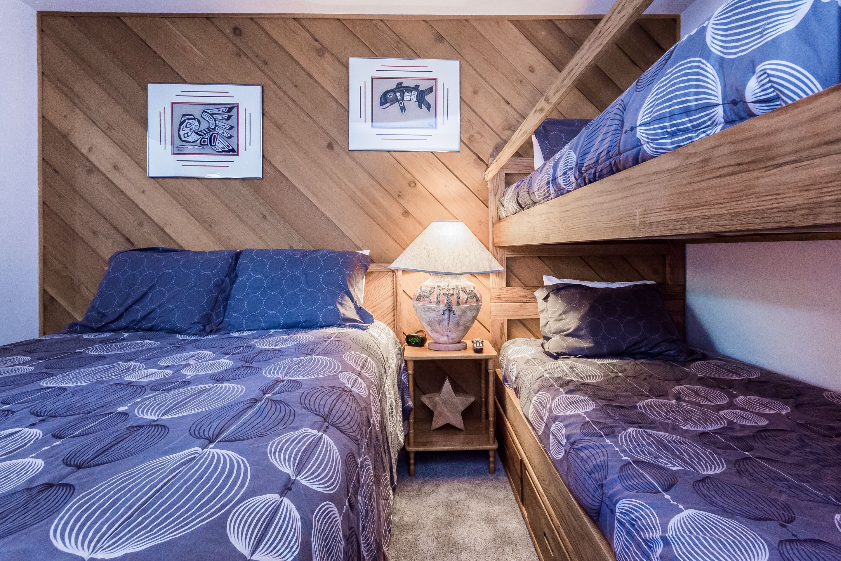 A bedroom with two bunk beds, a bedside table with a lamp, and framed artwork on the wall.