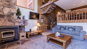 Ein gemütlicher Wohnraum mit Kamin aus Stein, einer Holztreppe, einem grauen Sofa und einem Flachbildfernseher.
