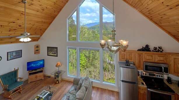 A spacious living room with a large window offering a mountain view, a wooden ceiling, a television, a sofa, and a kitchen area with modern appliances.