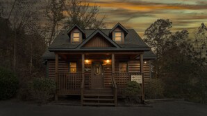 A rustic log cabin with a front porch, two windows, and a sign indicating it is a romantic getaway.
