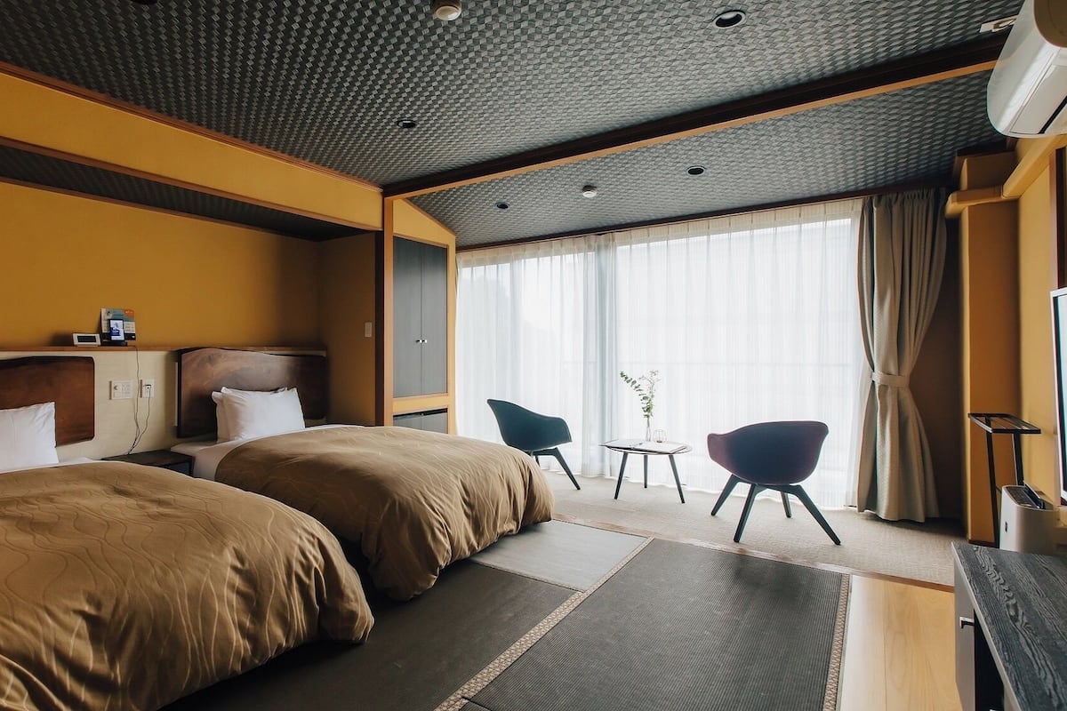 A hotel room with two beds, a desk, a chair, a TV, and a large window with curtains.