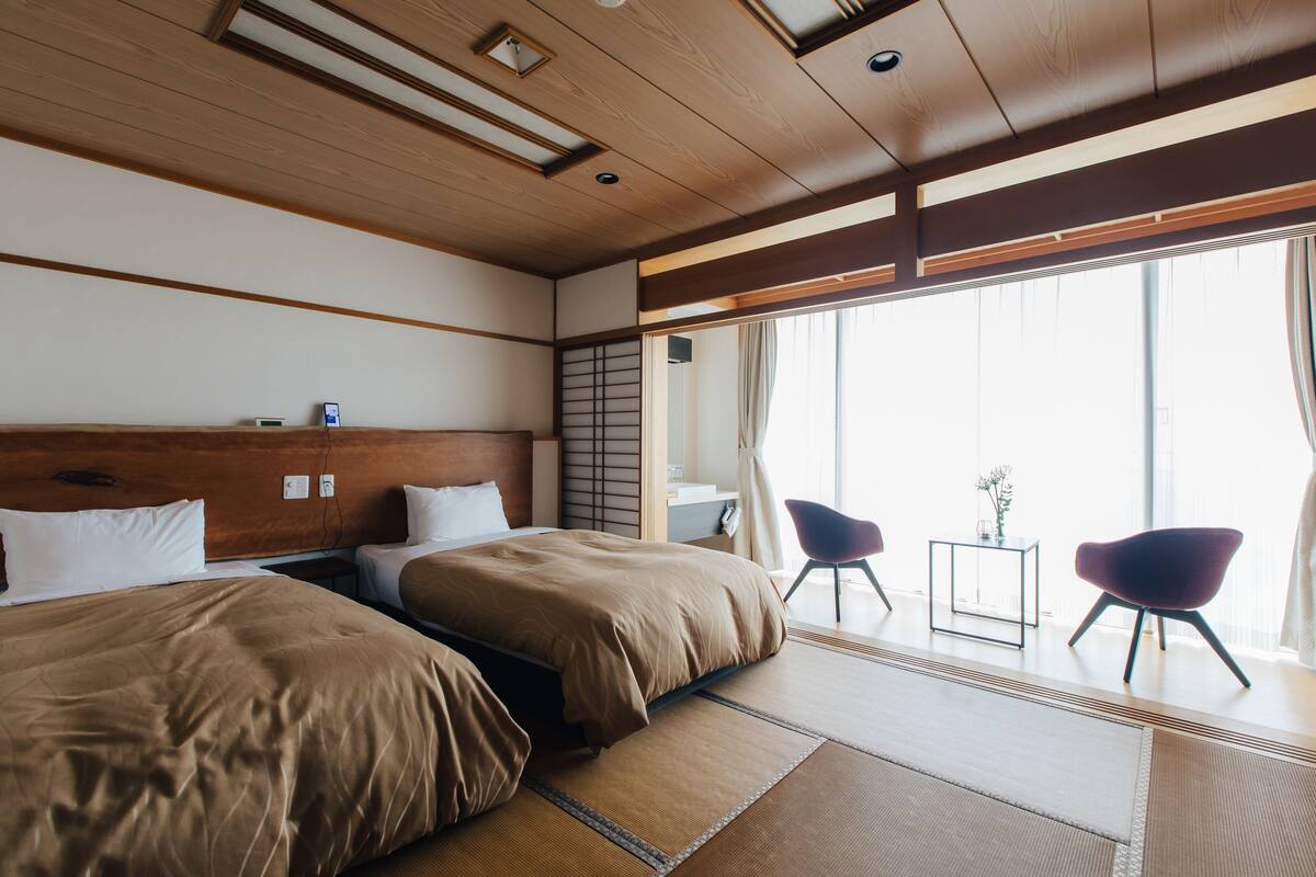 A hotel room with two beds, a small table, chairs, and a view of the outside.