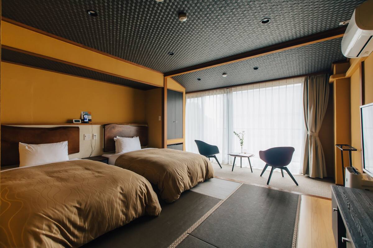 A hotel room with two beds, a desk, a chair, and a large window with curtains.