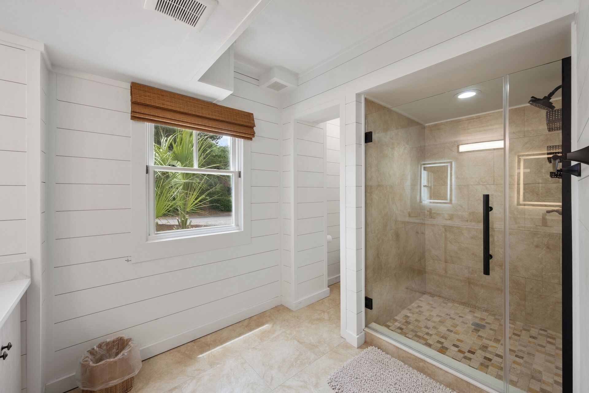 A bathroom with a glass-enclosed shower, a window with a view of greenery, and a white tiled floor.