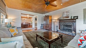 A spacious living room with a fireplace, a ceiling fan, and a dining area visible through an open door. - Second-Row 9BR Home | Ocean Views + Game Room Fun (Georgetown)