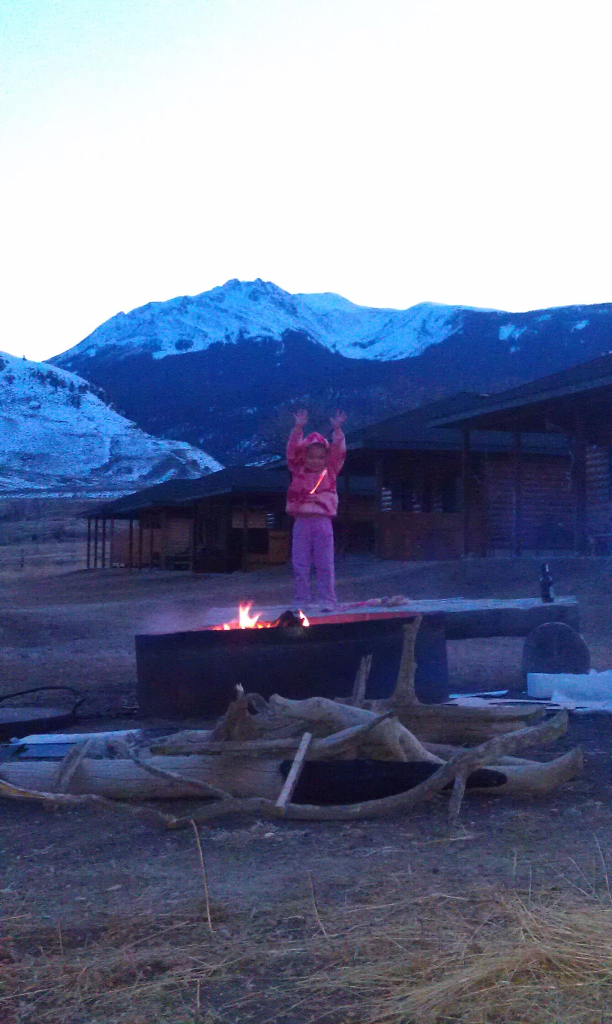Una persona de pie en un escenario con fuego al frente, rodeada por un entorno rústico al aire libre y montañas al fondo.