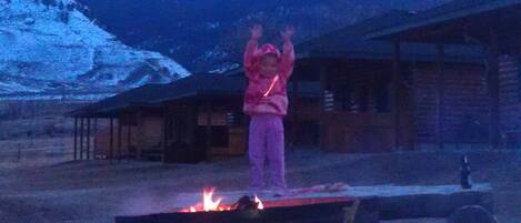 Una persona de pie en un escenario con fuego al frente, rodeada por un entorno rústico al aire libre y montañas al fondo.