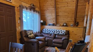 A cozy cabin interior with a brown leather sofa, a wooden rocking chair, and a dining table with a floral tablecloth.