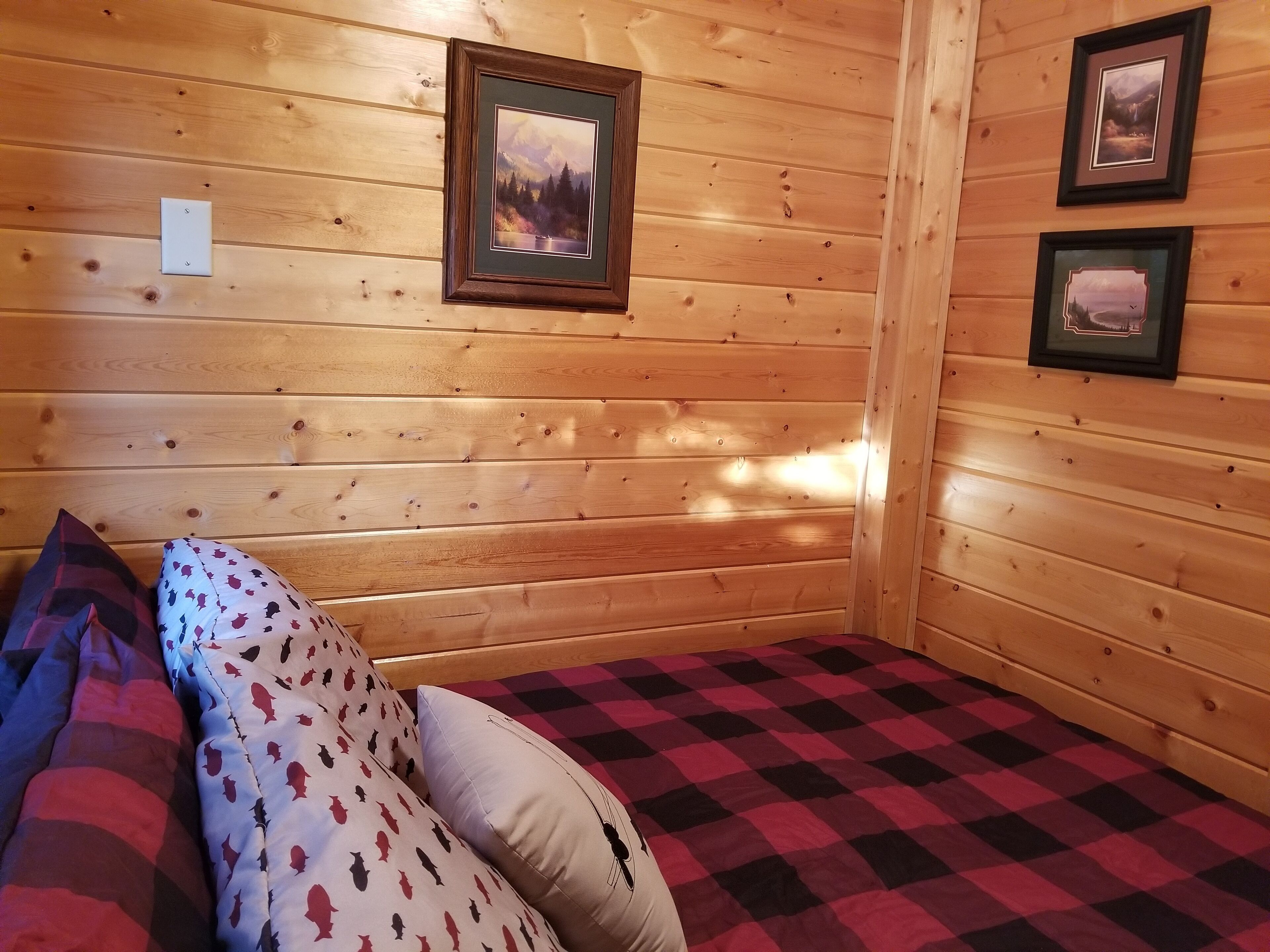 Une pièce chaleureuse aux murs en bois, avec une couvre-lit à carreaux et deux tableaux encadrés accrochés au mur.