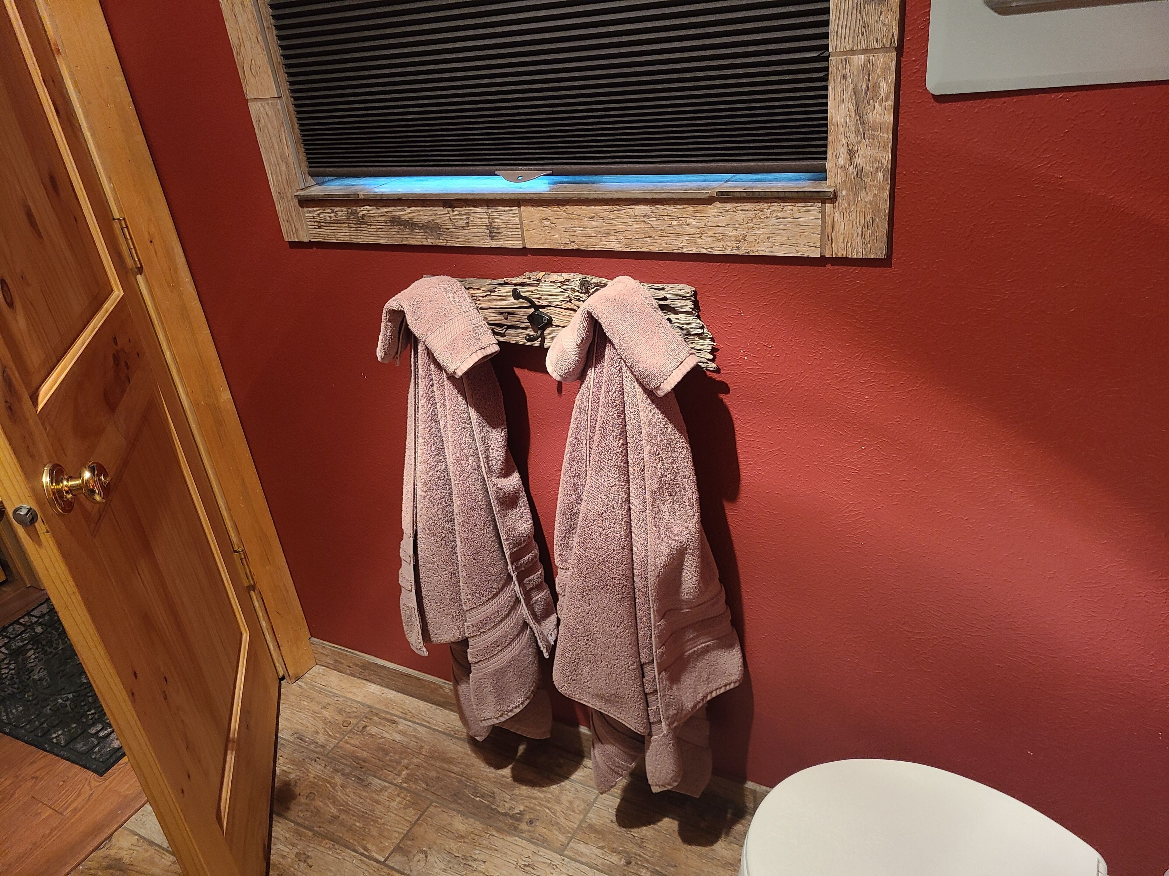 A bathroom with wooden flooring, a red wall, and a window with blinds.