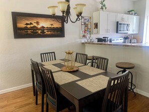 A dining area with a wooden table, six chairs, a chandelier, a framed picture, and a kitchen in the background.