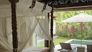 A four-poster bed with a canopy, a small table with flowers, and a view of a pool and garden. - Beautiful, Peaceful Villa/Excellent Location/Wonderful Balinese Staff (Penestanan, Ubud)