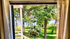 A table with a vase of flowers, a candle, and a tray with a glass of water, positioned by a window with a view of a park and a body of water.