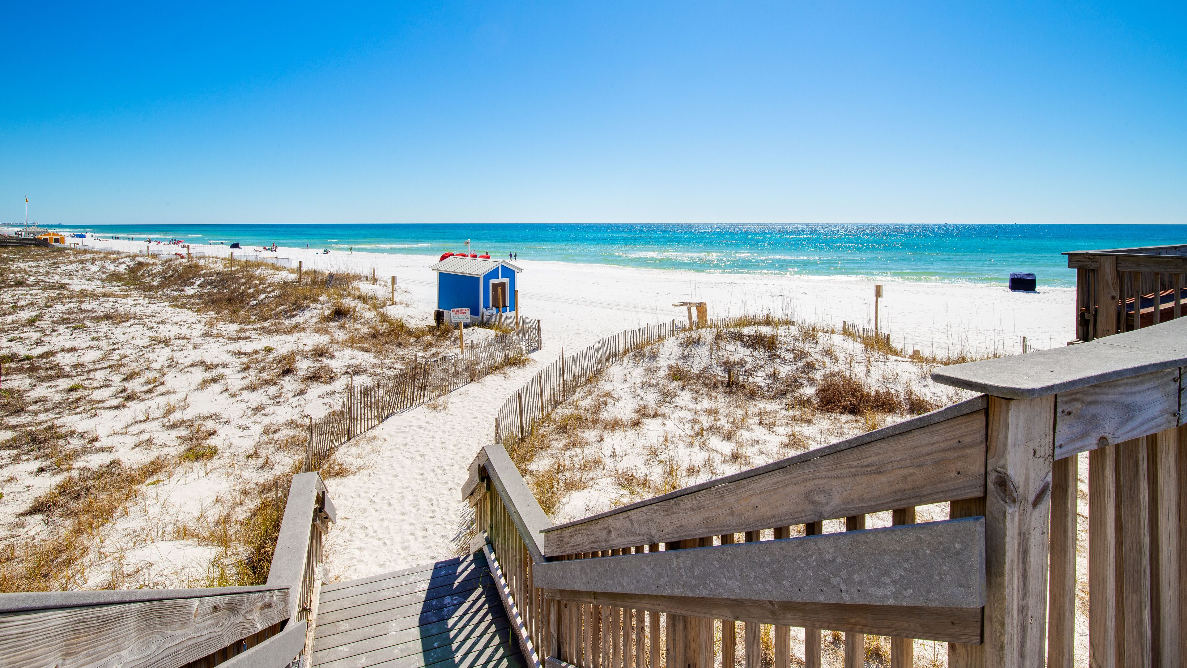 A beach with a wooden boardwalk leading to a small blue structure, sandy dunes, and a clear blue sky.