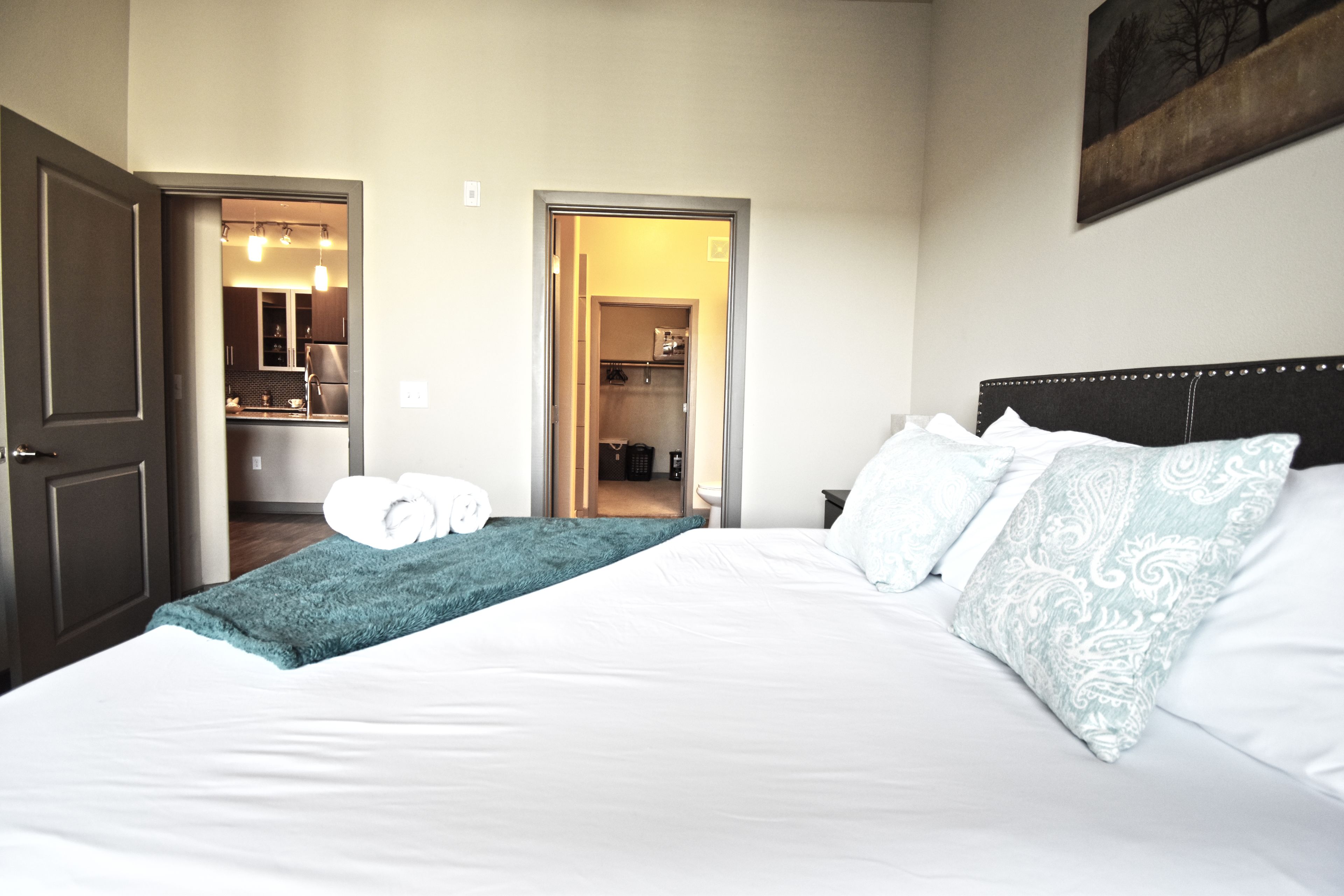 A hotel room with a large bed, a bedside table, a nightstand, and a view of the bathroom and kitchen through an open door.