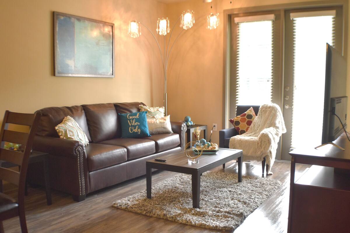 A living room with a brown leather sofa, a coffee table, and a white armchair. There is a painting on the wall and a floor lamp.