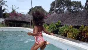 A child jumping into a swimming pool with a thatched-roof structure in the background. - Travellers Inn Resort Malindi (Malindi)