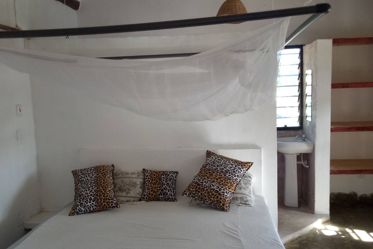 Una cama con sábanas blancas y almohadas de estampado de leopardo, un ventilador de techo y una ventana con luz natural.