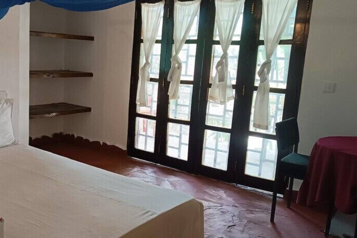 Una habitación con cama, ventana con cortinas, una silla y una mesa con mantel rojo.