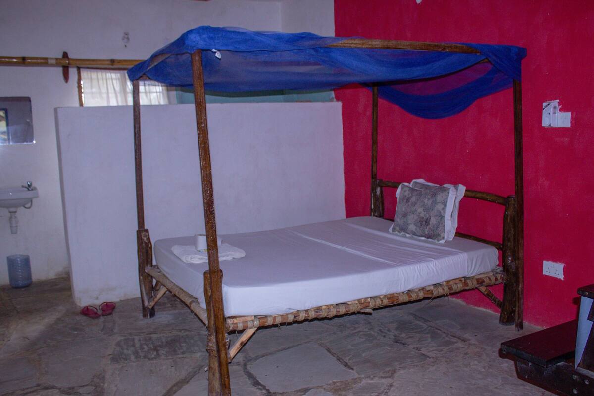 Una cama individual con dosel azul, cabecera de madera y almohada. Se ve un lavabo blanco y una botella de agua azul en la habitación.