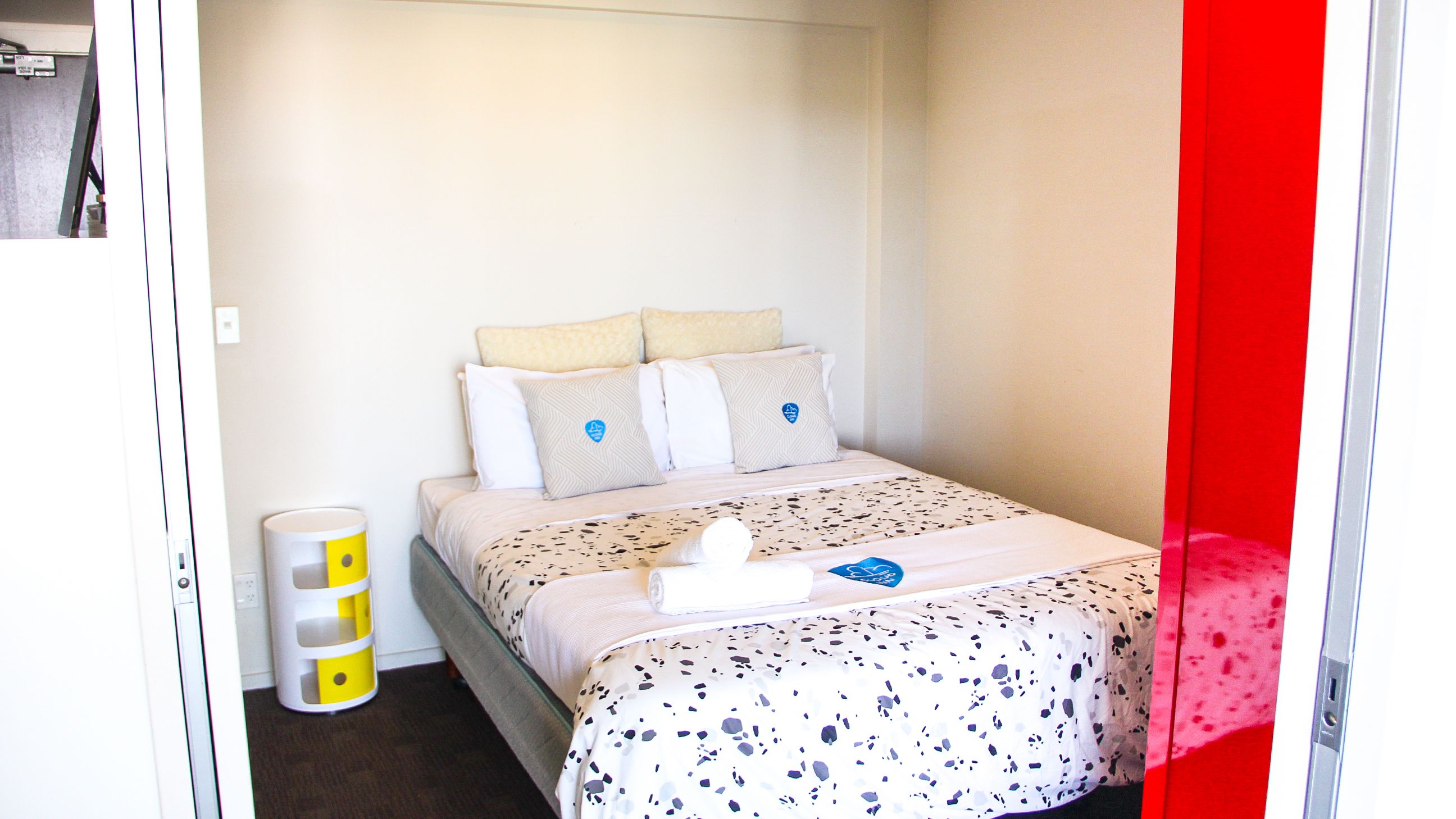A small, neatly arranged bedroom with a bed, pillows, a bedside table, and a trash bin.