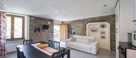 A dining area with a table and chairs, a sofa, a stone wall, a television mounted on the wall, and a kitchen area with a microwave.