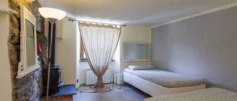 A small, cozy room with a stone wall, a bed, a bedside table, a lamp, and a window with curtains.