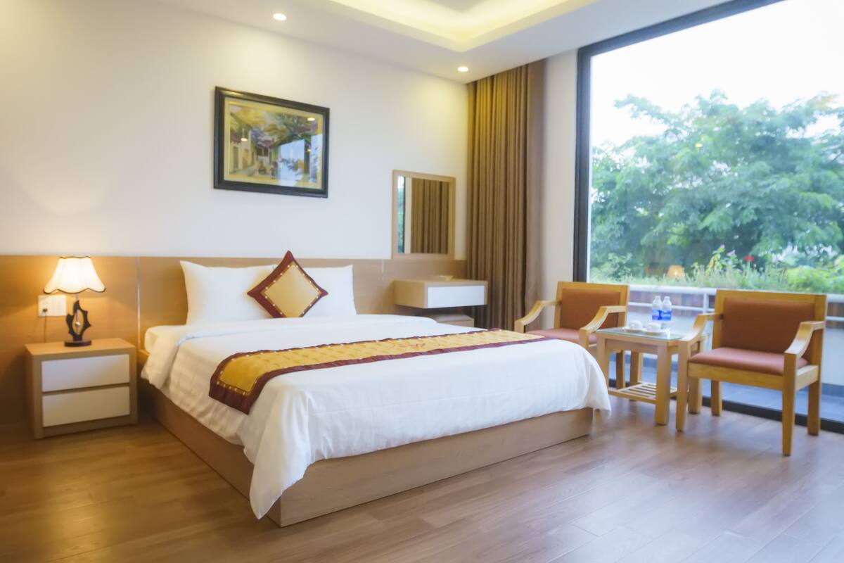 A hotel room with a bed, bedside table, lamp, chair, and a view of greenery through a large window.