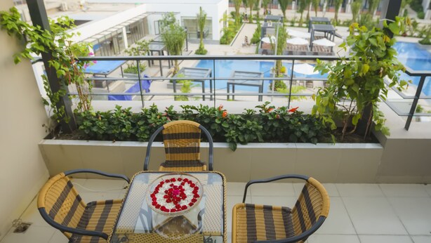 Un balcón con sillas de mimbre y una mesa, con vistas a una zona de piscina con sillones de descanso y vegetación.