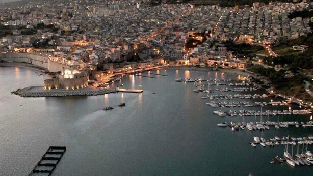 A coastal cityscape with a marina filled with boats, a prominent waterfront promenade, and densely packed buildings.