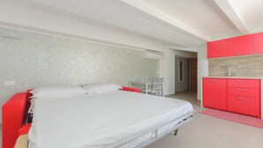 A bedroom with a bed, a small table, and a kitchenette area with red cabinets.