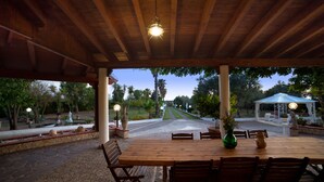 Une terrasse extérieure avec une table et des chaises en bois, une pergola agrémentée d’une suspension lumineuse, et une vue sur un jardin et un sentier.