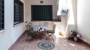 Un pequeño patio con una mesa redonda y sillas, una ventana con rejas y suelo de baldosas.