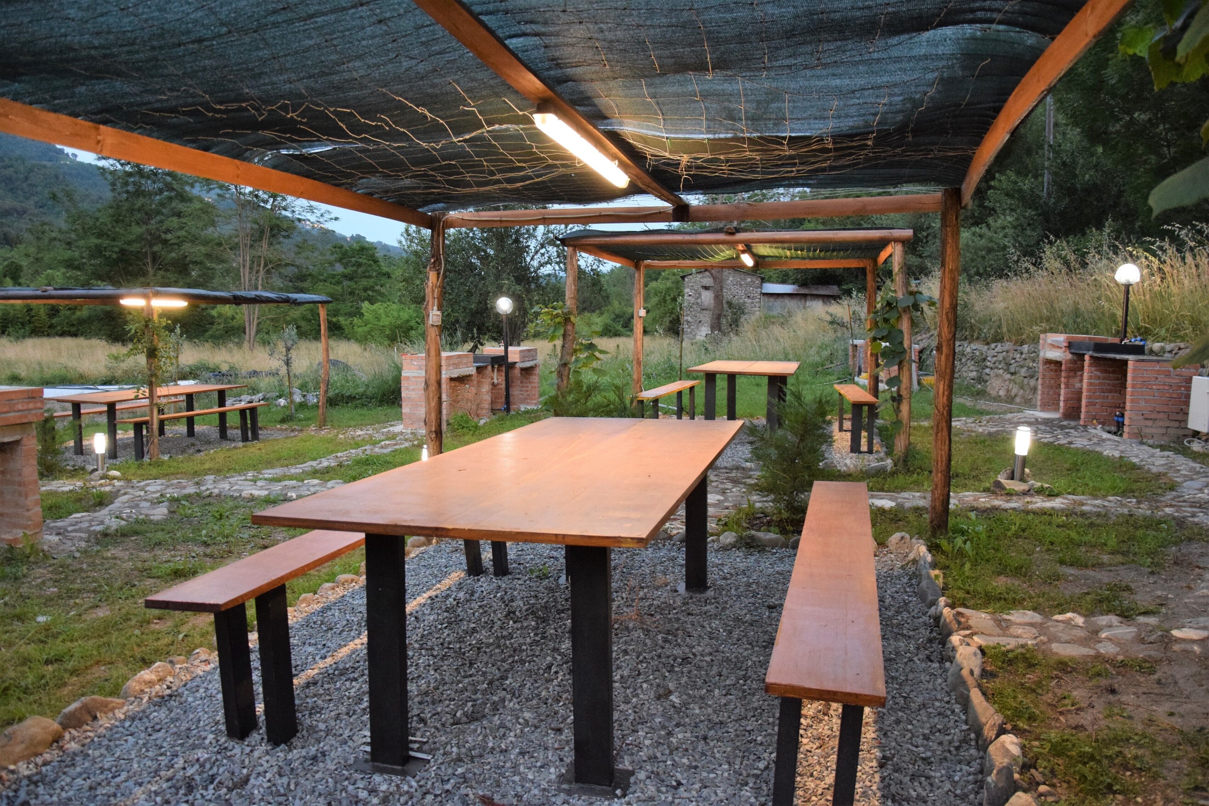 An outdoor patio area with wooden tables and benches, surrounded by a gravel path and greenery.