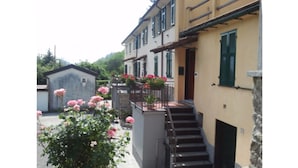 A building with a staircase leading to a second floor, surrounded by greenery and pink flowers. - Casa Rosa "Casa Stefano" (Pian dei Manzi)