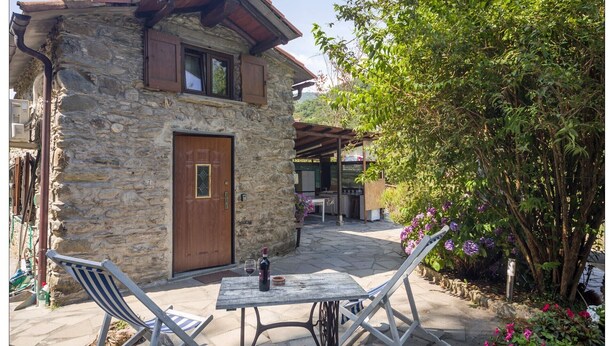 A stone building with a wooden door, a covered outdoor seating area, and a garden with purple flowers. - Casa Rosa "Casa Stefano" (Pian dei Manzi)