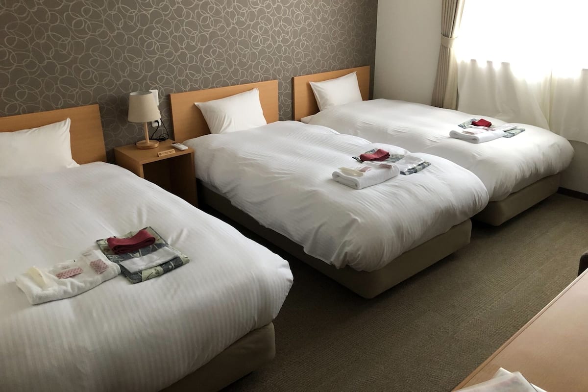 A hotel room with two beds, each with white linens and towels. There is a nightstand with a lamp and a remote control. The room has a patterned wallpaper and a window with curtains.