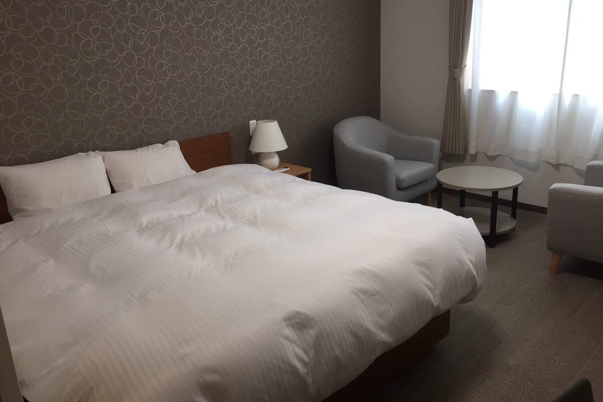 A hotel room with a large bed, a bedside table with a lamp, a grey armchair, a small round table, and a patterned wall.