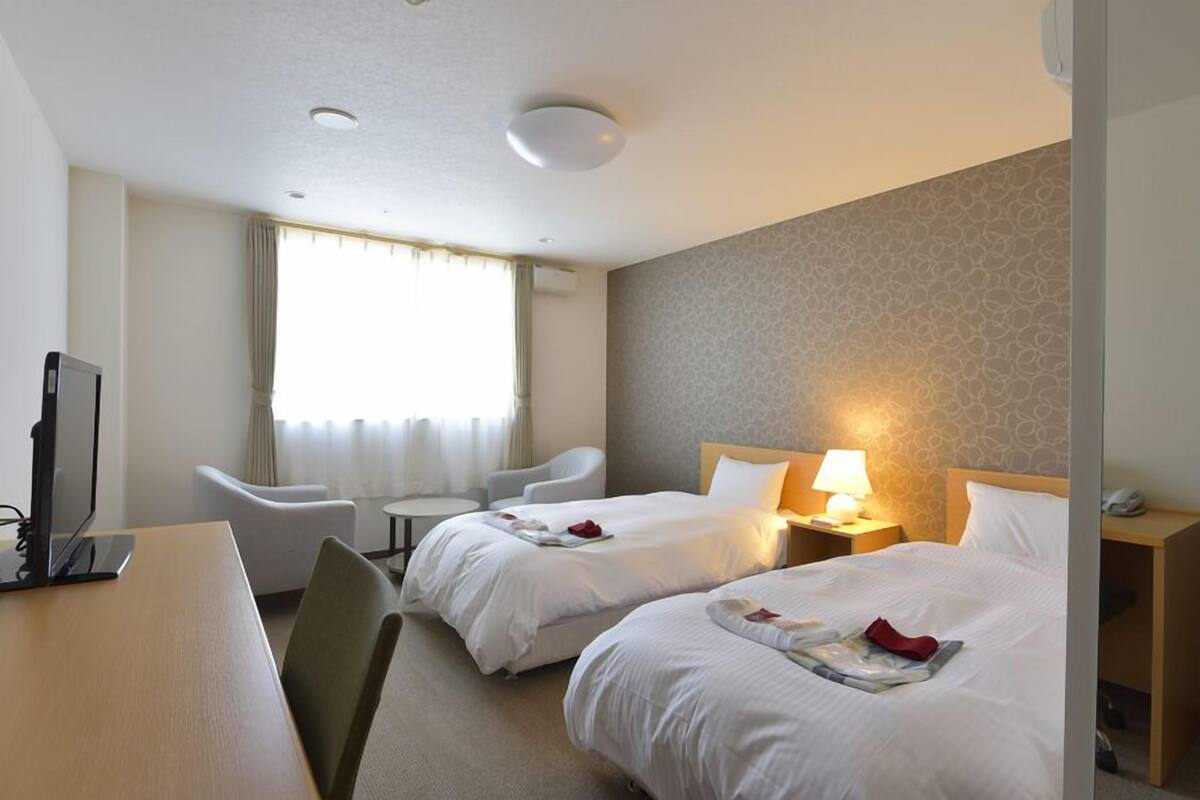 A hotel room with two beds, a desk, a chair, a lamp, and a TV.