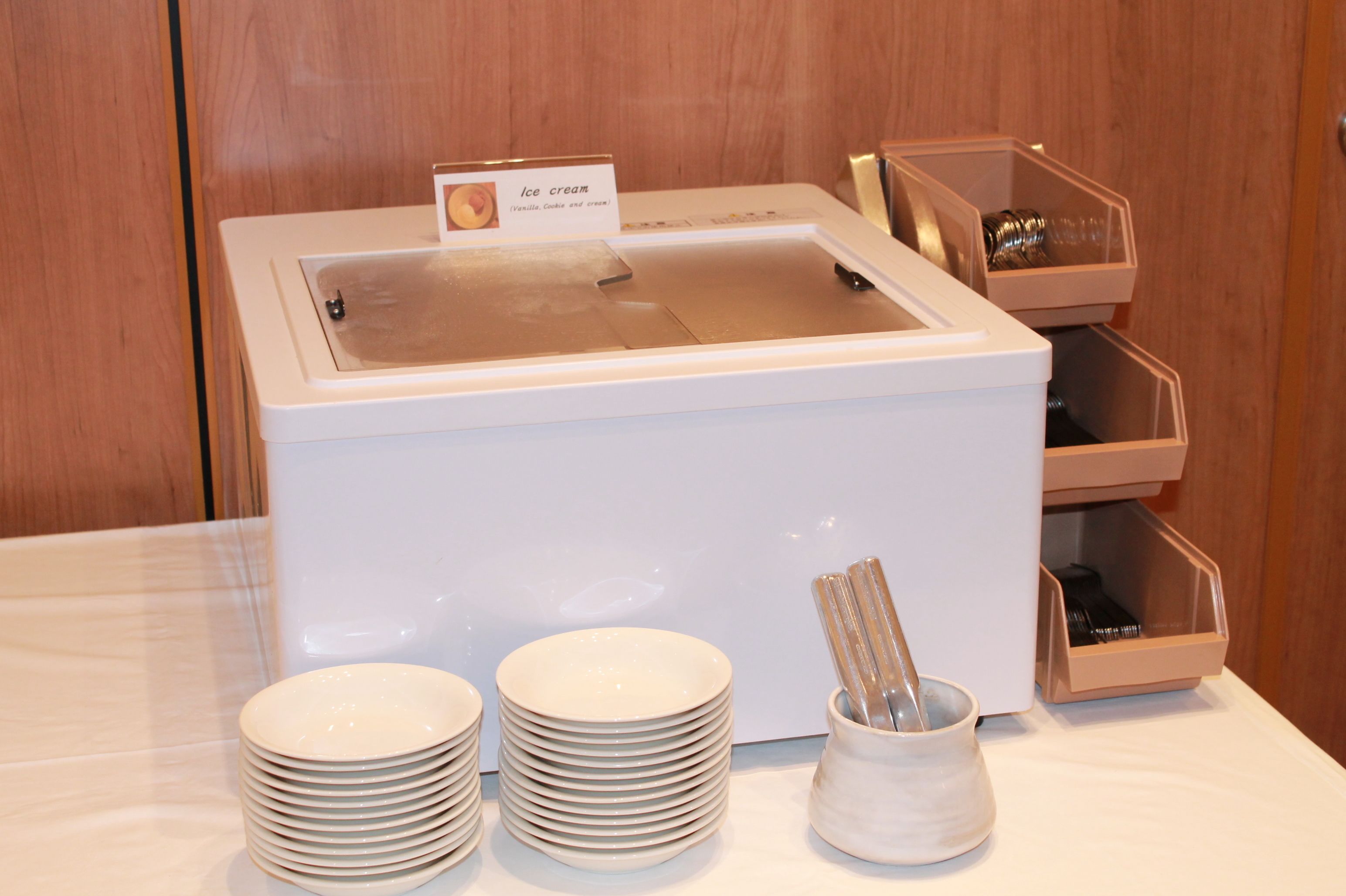 A buffet setup with a sign indicating ice cream, a stack of white plates, and a utensil holder.