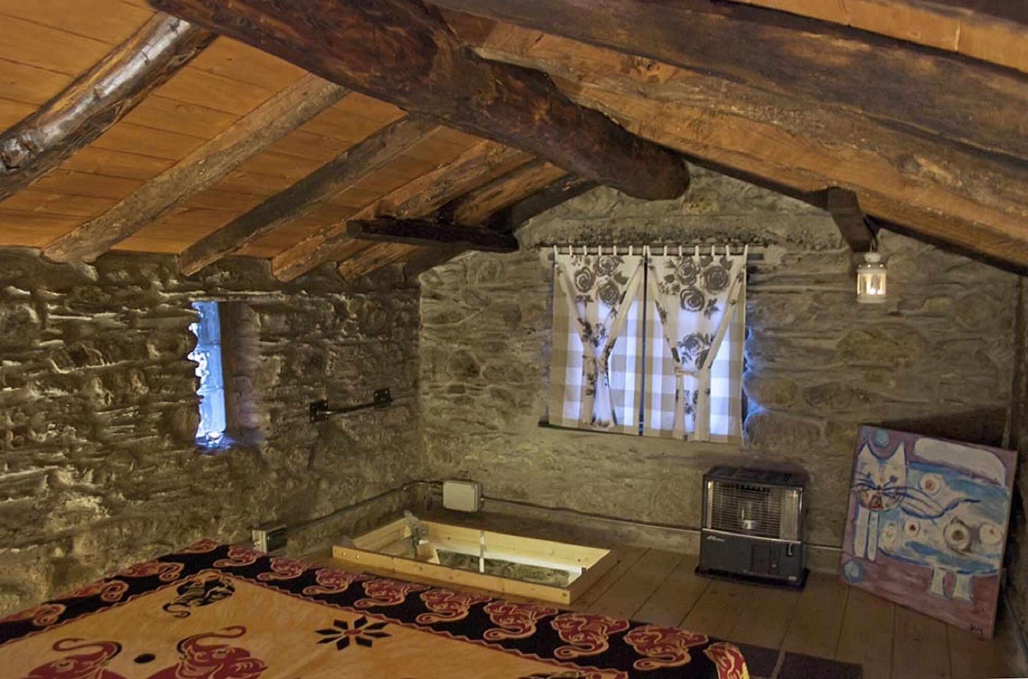 Une chambre mansardée au style rustique, avec des poutres apparentes, un mur en pierre, une fenêtre ornée de rideaux à motifs floraux, un radiateur et un tableau coloré.