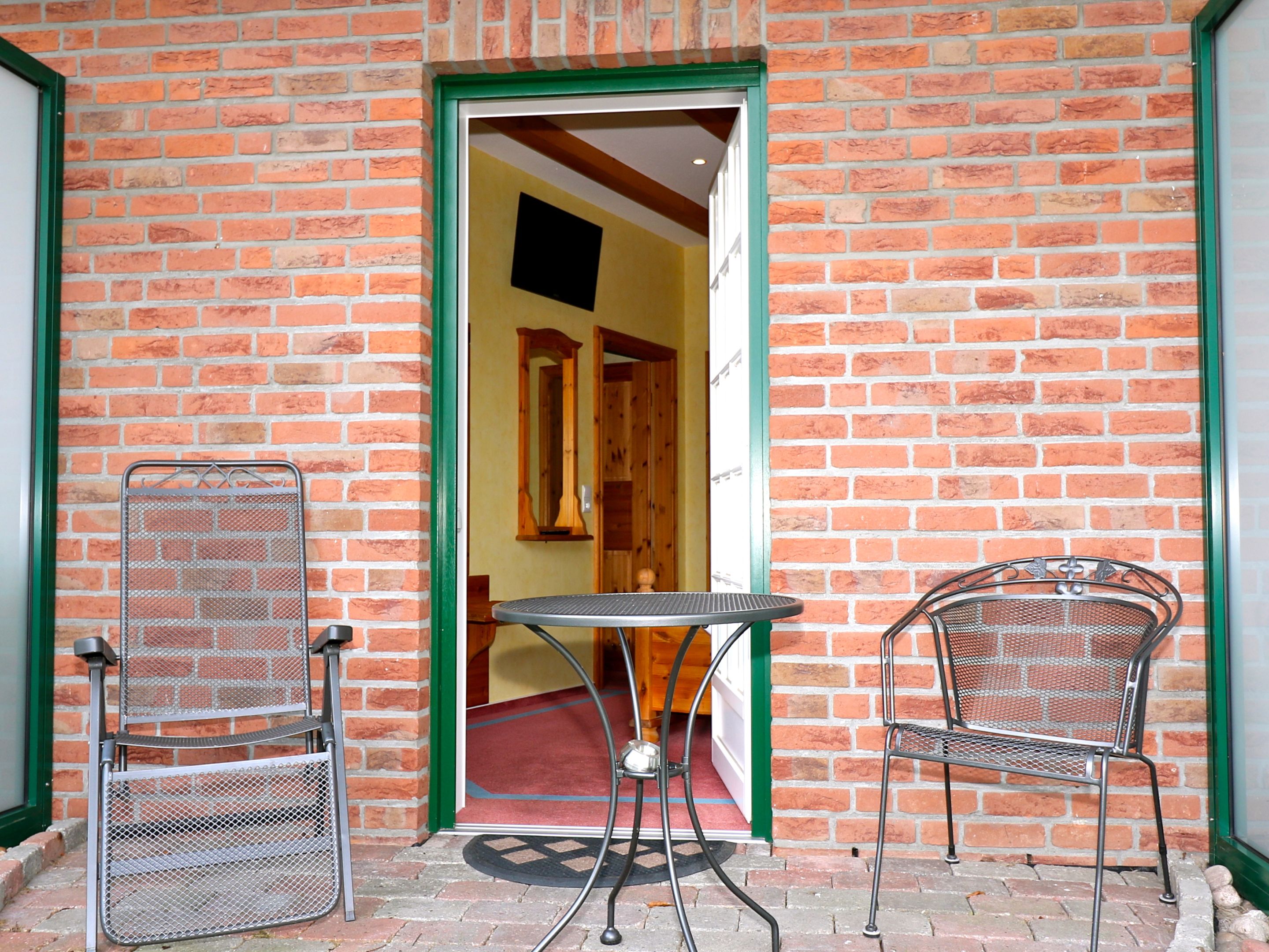 Ein Backsteinhaus mit einer grün gerahmten Tür, die zu einem Innenraum mit einer Holztür und einem Spiegel führt. Vor der Tür steht ein Metalltisch und zwei Metallstühle.