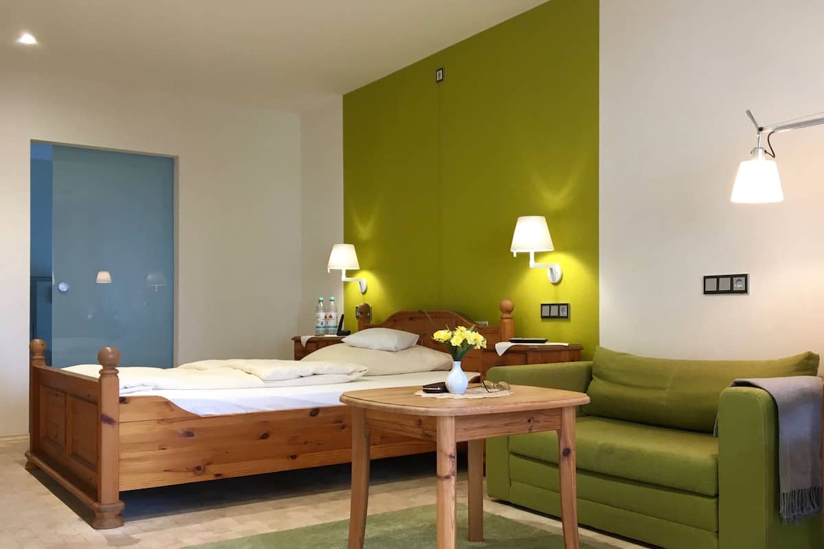 Habitación de hotel con una cama de madera, paredes verdes, dos lámparas de noche, un sofá verde y una pequeña mesa de madera.