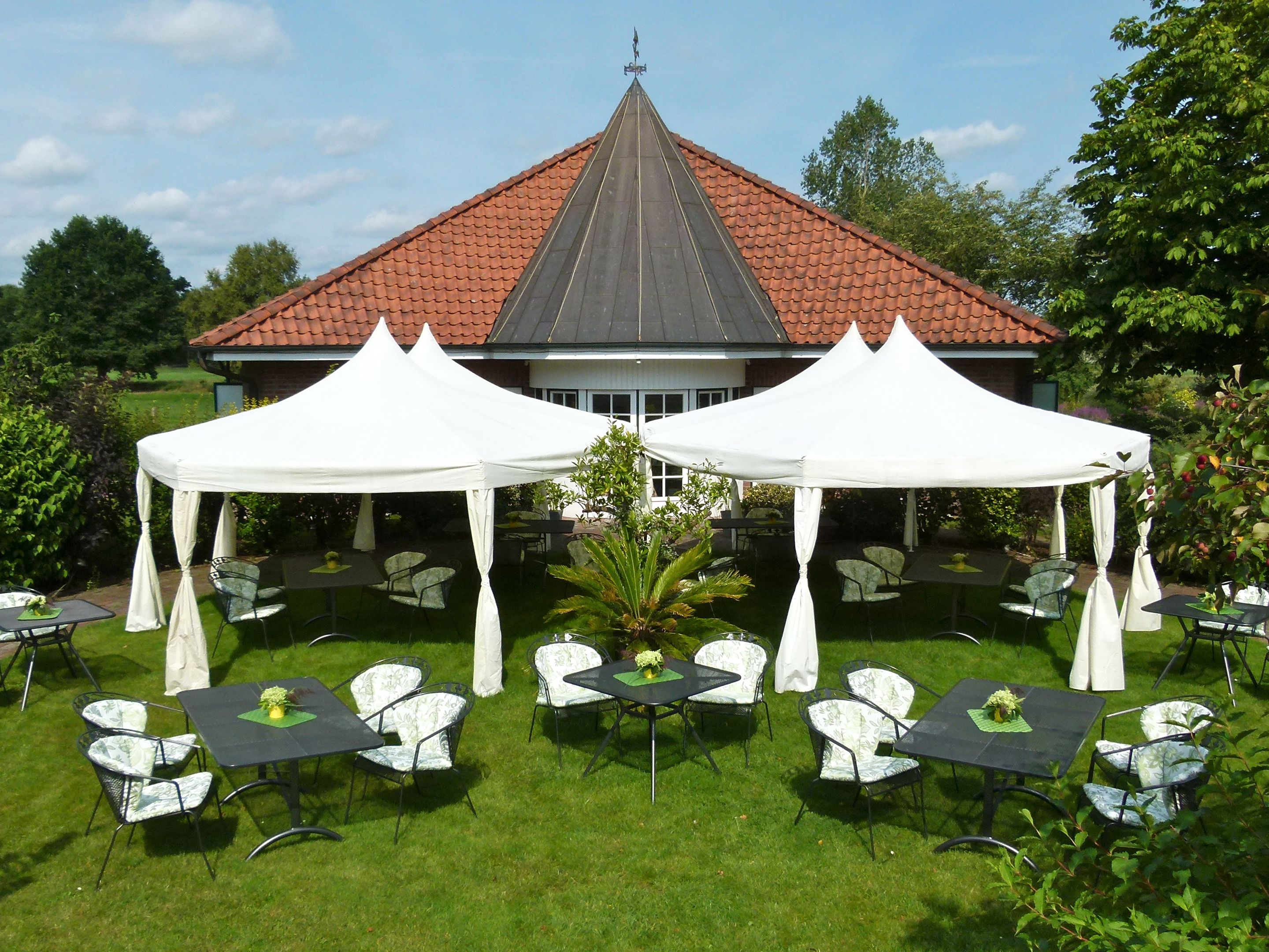 Ein Außenbereich mit Terrassenmöbeln, einer weißen Zelthalle und einem Gebäude mit rotem Dach im Hintergrund.